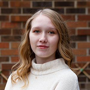 A headshot of a girl with blonde hair and a white sweater smiling at the camera in front of a brick wall